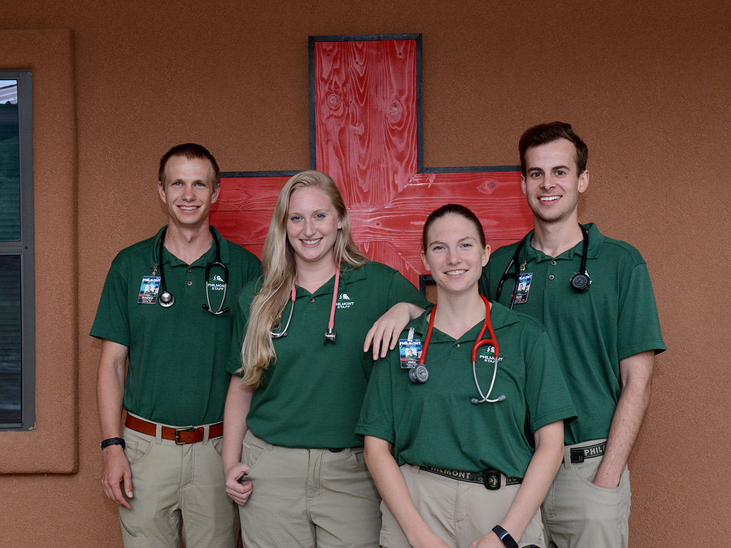 Four students posing in front of a red medical cross with stethoscopes around their necks